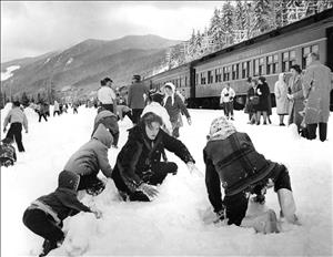 Children in winter clothes play in the snow while adults stand beside a passenger train in the mountains. 